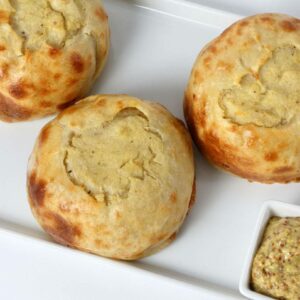 Seven handmade baked potato knishes on a white tray, served with a small square dish of whole grain mustard on the side.