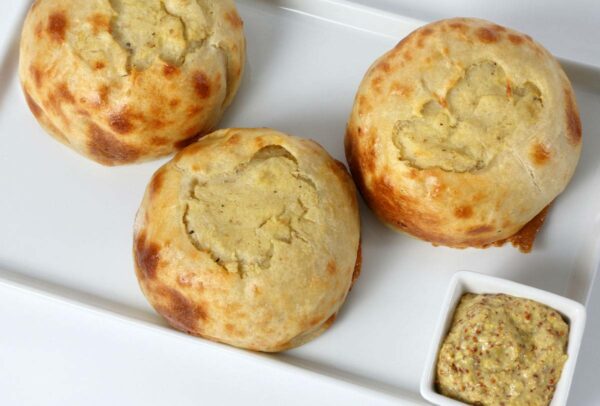 Seven handmade baked potato knishes on a white tray, served with a small square dish of whole grain mustard on the side.