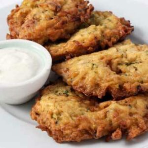 Four zucchini pancakes, served on a white plate with a small bowl of white dipping sauce.