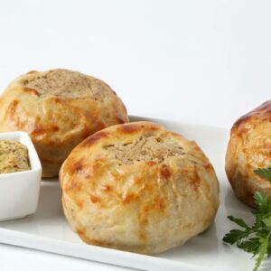Seven handmade baked kasha knishes are arranged on a white plate, served with a small dish of grainy mustard and a sprig of fresh parsley.