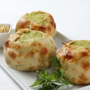 Three Handmade Baked Broccoli Knishes are served on a rectangular white plate, accompanied by grainy mustard in a small bowl and a fresh herb sprig.