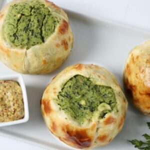 Seven handmade baked spinach knishes are arranged on a white tray beside a small dish of whole grain mustard and a sprig of parsley.